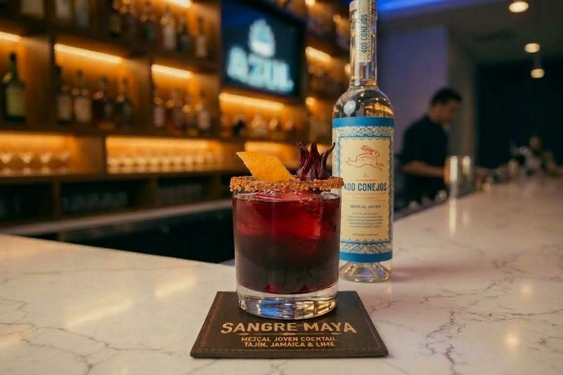 A Sangre Maya cocktail with a hibiscus garnish and an orange peel beside a bottle of 400 Conejos mezcal, with lit bottle shelves behind